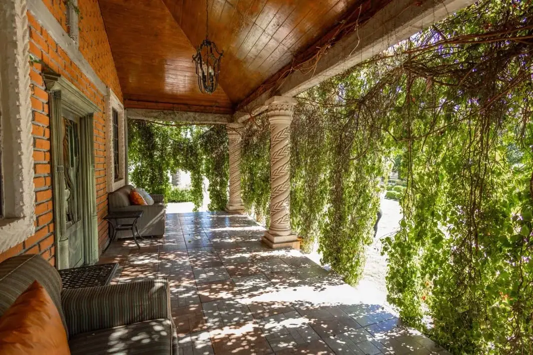 Corredor con columnas, techo de madera y enredadera, con sillones a la sombra
