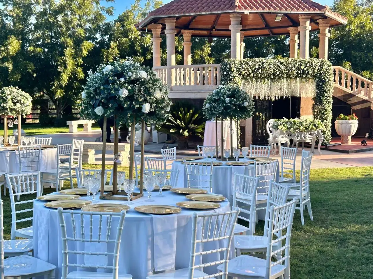 Mesas redondas con mantel blanco, sillas blancas y centros de mesa florales frente al kiosko decorado