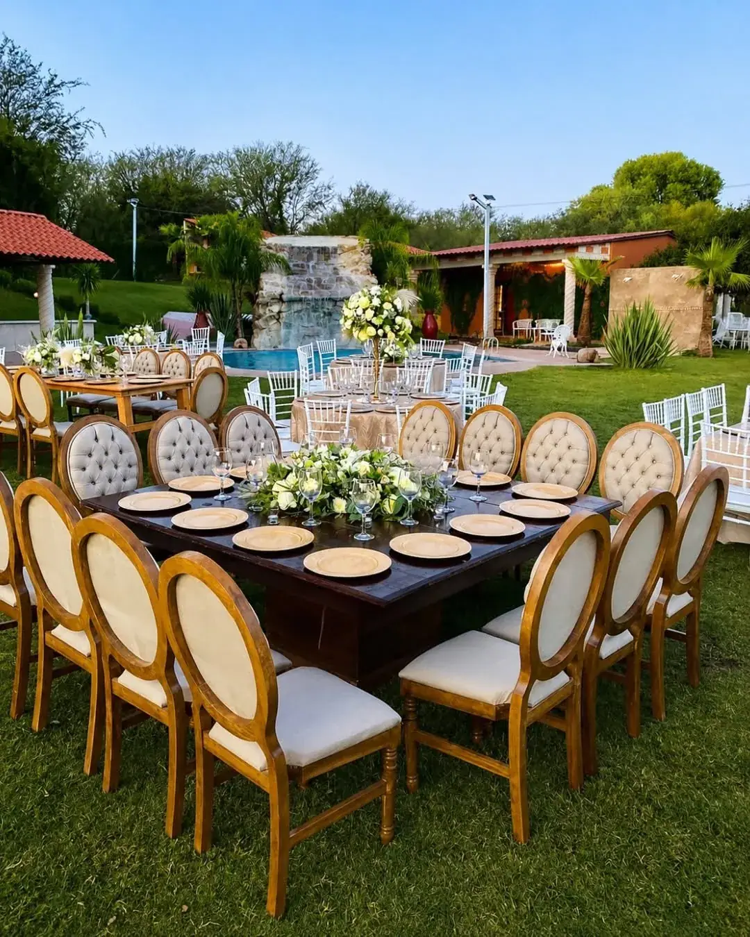 Mesa imperial de madera con sillas tapizadas, platos dorados y flores blancas en el jardín