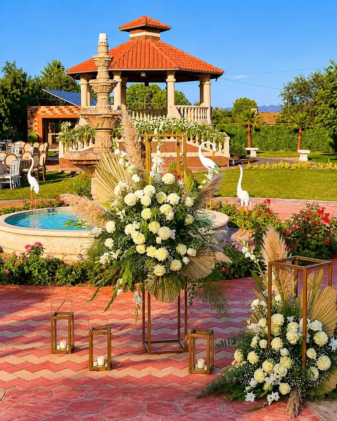 Arreglo de flores blancas y faroles dorados junto a la fuente, con el kiosko al fondo